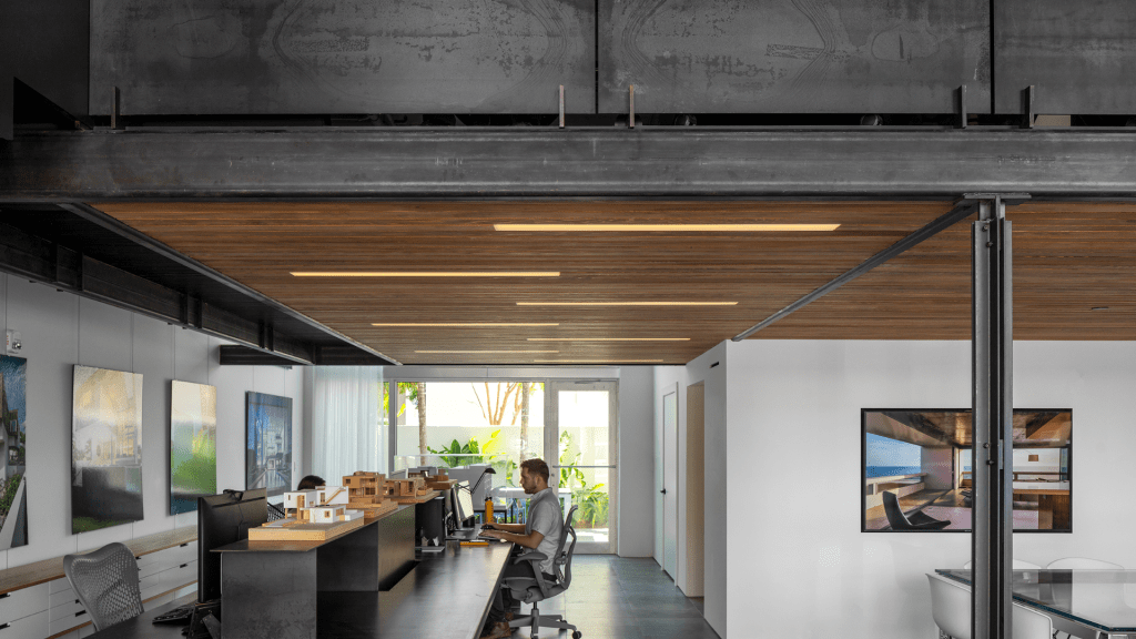 Modern office with two people working at desks, large windows in the back letting in natural light, wood ceiling panels, white walls, and framed photos of architecture on the walls.