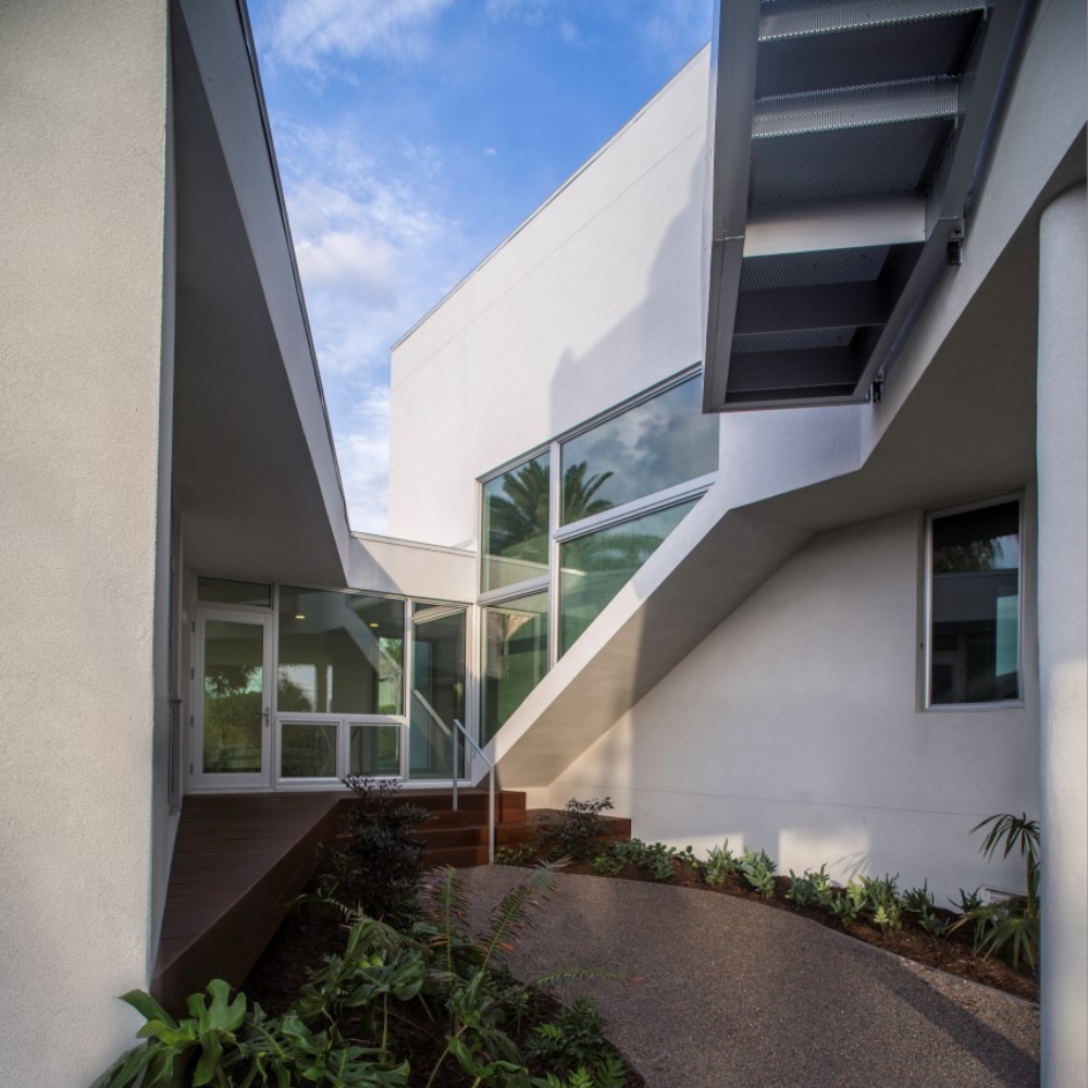 Modern white building with clean lines, large windows, and an exterior staircase. Curved walkway bordered by plants leads to the entrance. Blue sky and palm tree reflections visible in the glass.