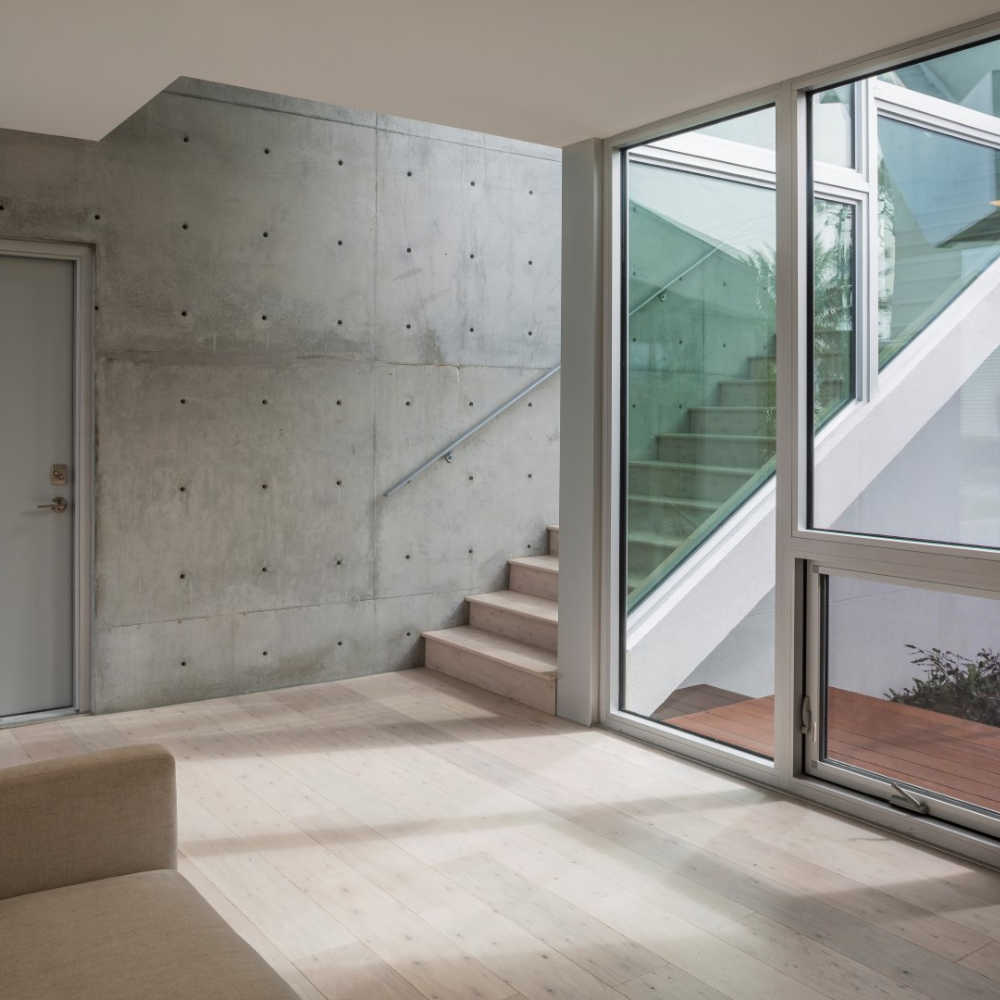 Modern, minimalist interior with concrete wall, light wood floor, and beige sofa. Stairs lead up beside a large window, revealing a glass-paneled outdoor staircase with sunlight streaming in.