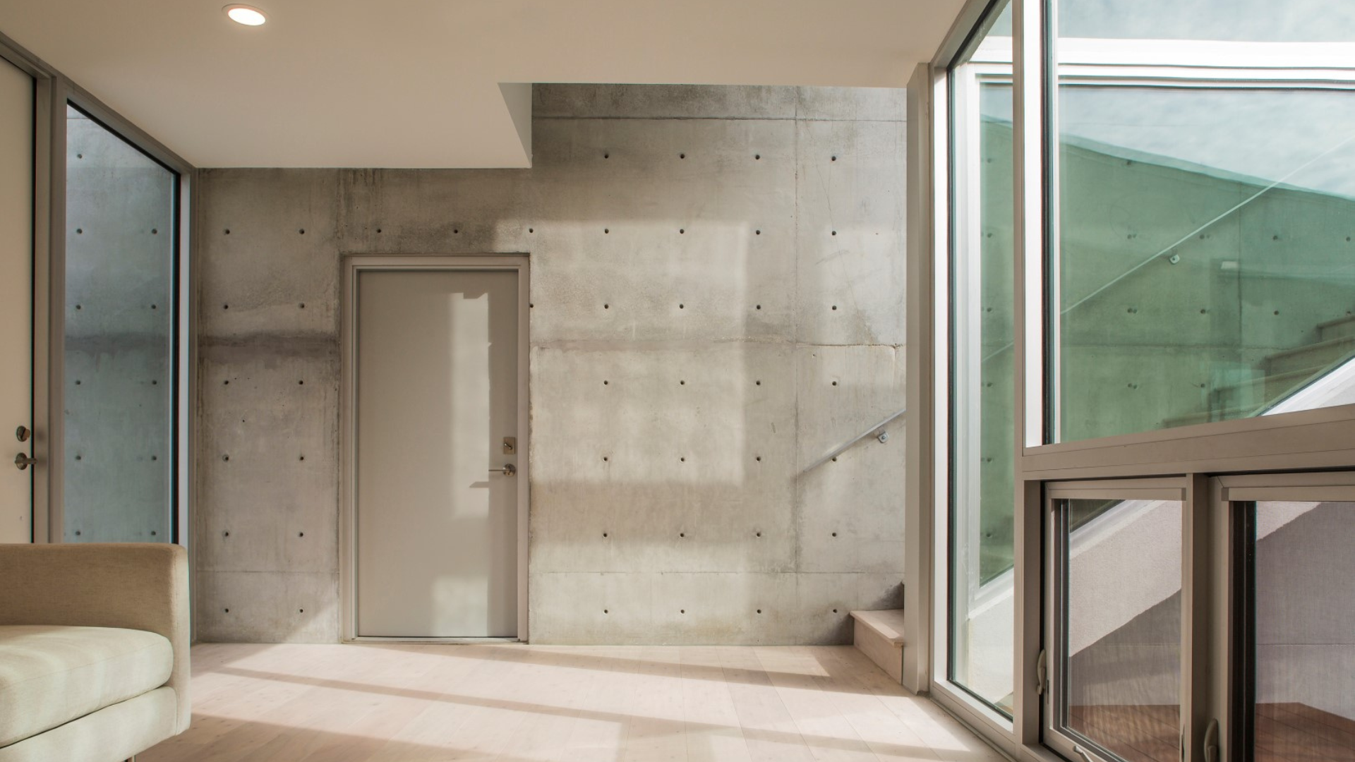 A modern interior with large glass windows, a concrete wall with a closed gray door, sunlight streaming in, and stairs leading up on the right side.