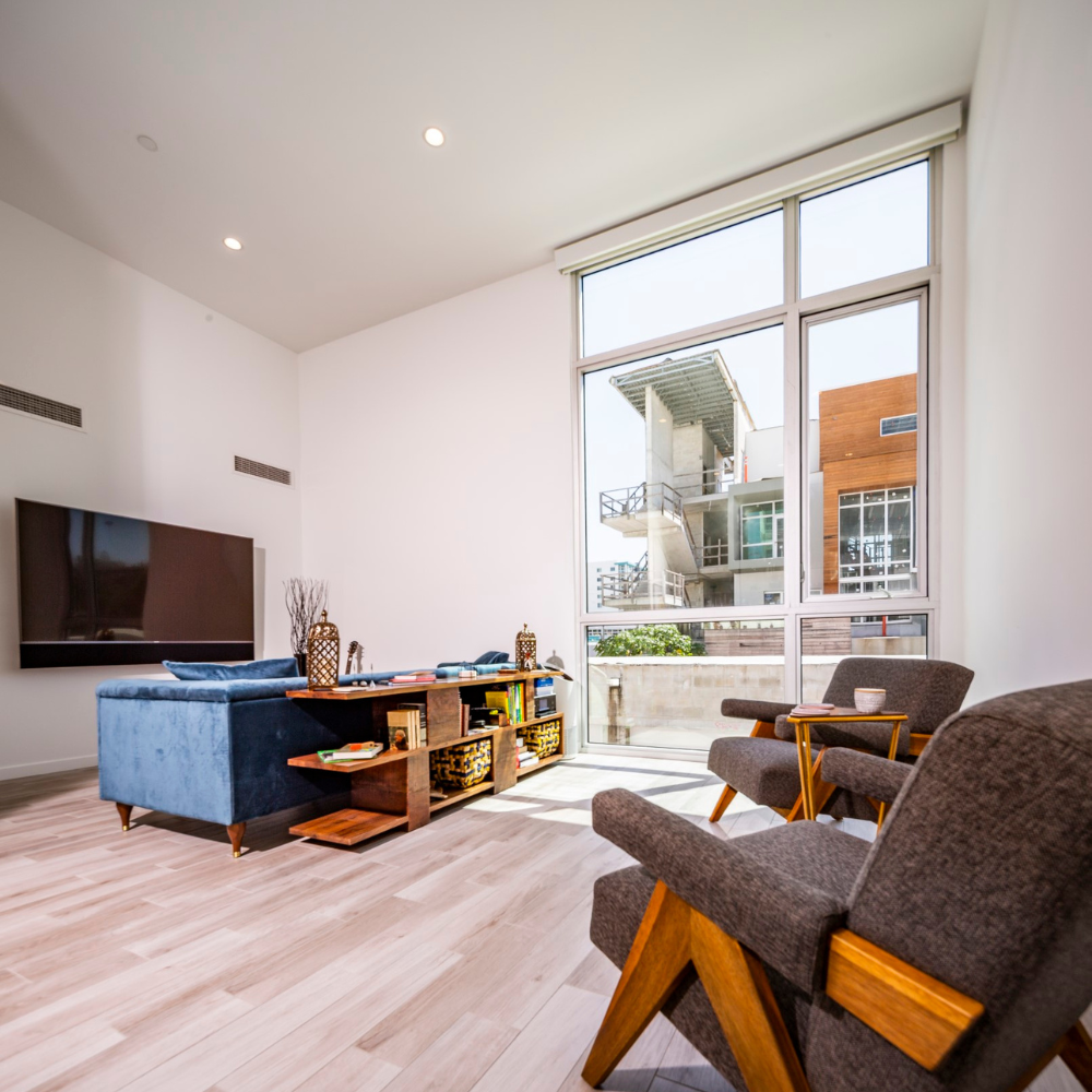 Modern living room with large windows, a blue sofa, two gray chairs, a wooden coffee table, and a wall-mounted TV. Bright natural light fills the minimalist space with light wood flooring.