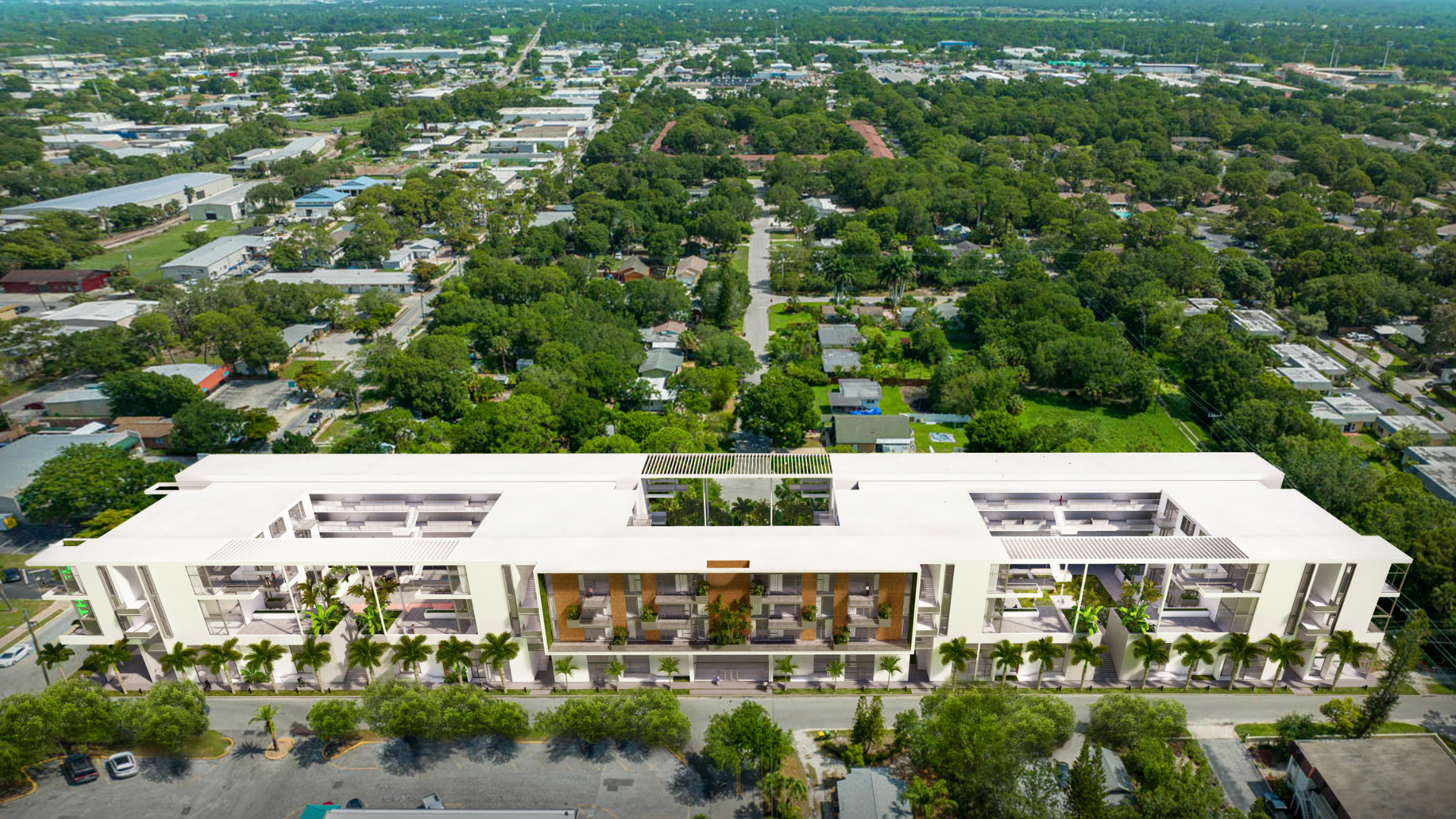 Aerial view of a modern, white, multi-story building lined with palm trees, surrounded by a green, tree-filled suburban neighborhood with houses and roads extending into the distance.