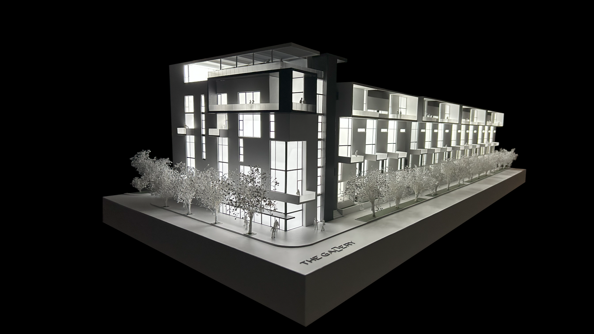 A white architectural model of a modern multi-story building with large windows, lined with small trees, displayed on a rectangular base labeled THE GALLERY against a black background.