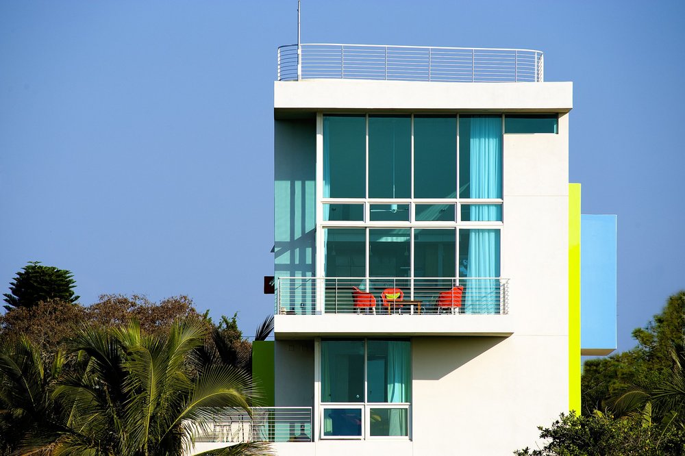 Modern white building with large blue-tinted windows and balconies, surrounded by palm trees. This award-winning property features four red chairs and a table on the balcony under a clear blue sky.