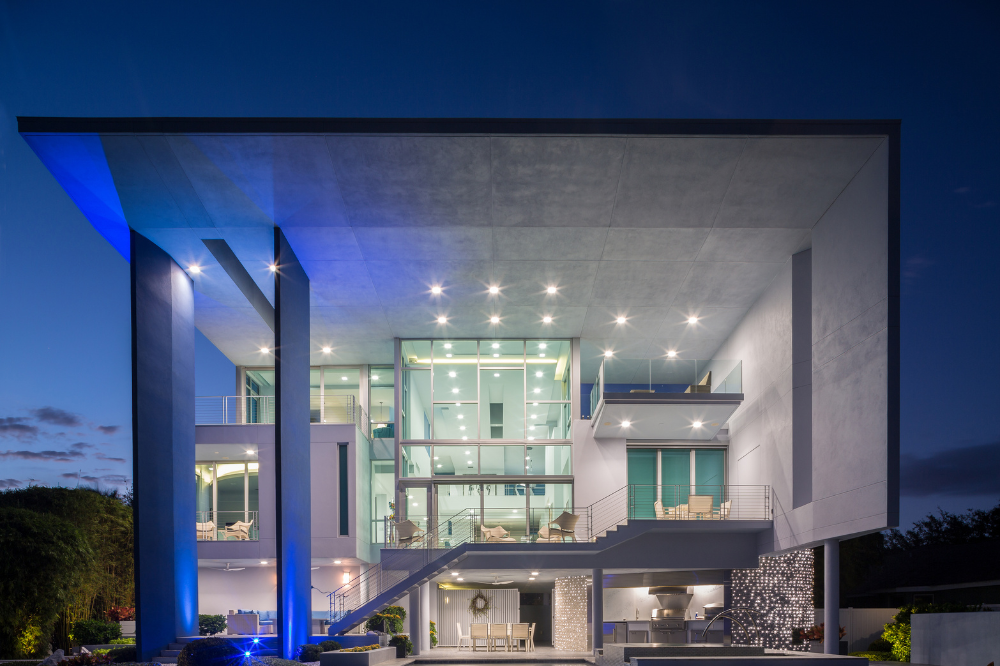 Modern, multi-story house with large glass windows, outdoor balconies, and bright lights illuminating the award-winning structure at dusk. The architecture features bold lines, tall pillars, and a spacious terrace area.