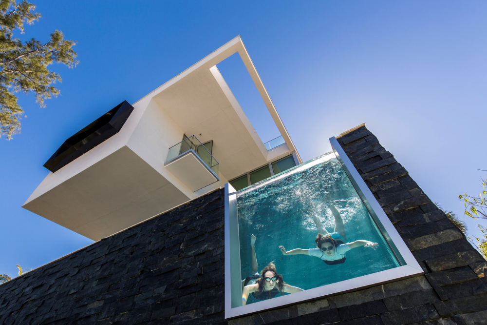 Two people swim in a clear pool with a glass wall, attached to an award-winning modern house with bold, angular architecture, viewed from below against a blue sky.