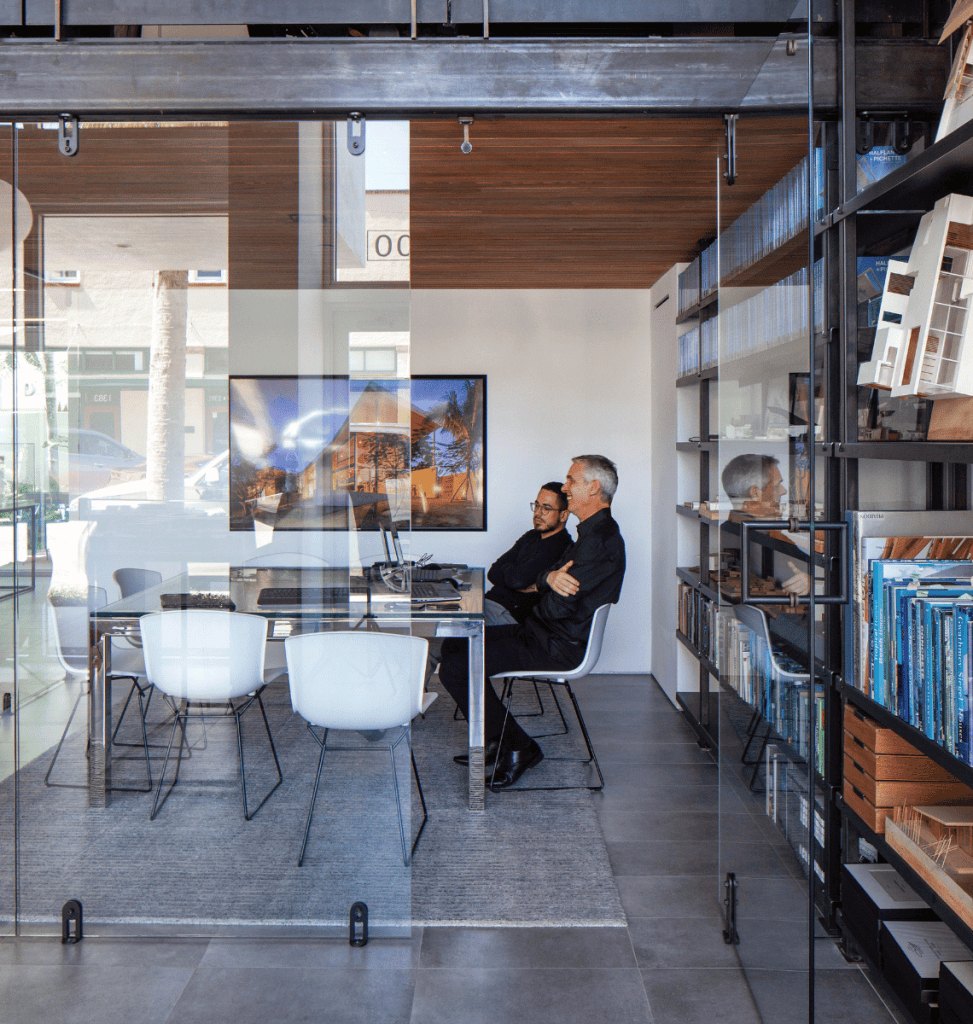 Two men sit at a glass table in a modern office, surrounded by shelves of books and a framed photo—natural light pours in, cars passing outside as if each holds its own story.