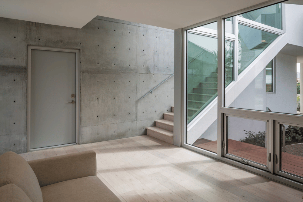 A modern interior with a light wood floor, concrete wall, gray door, and large windows showing a staircase and natural light streaming in—an award-worthy design.