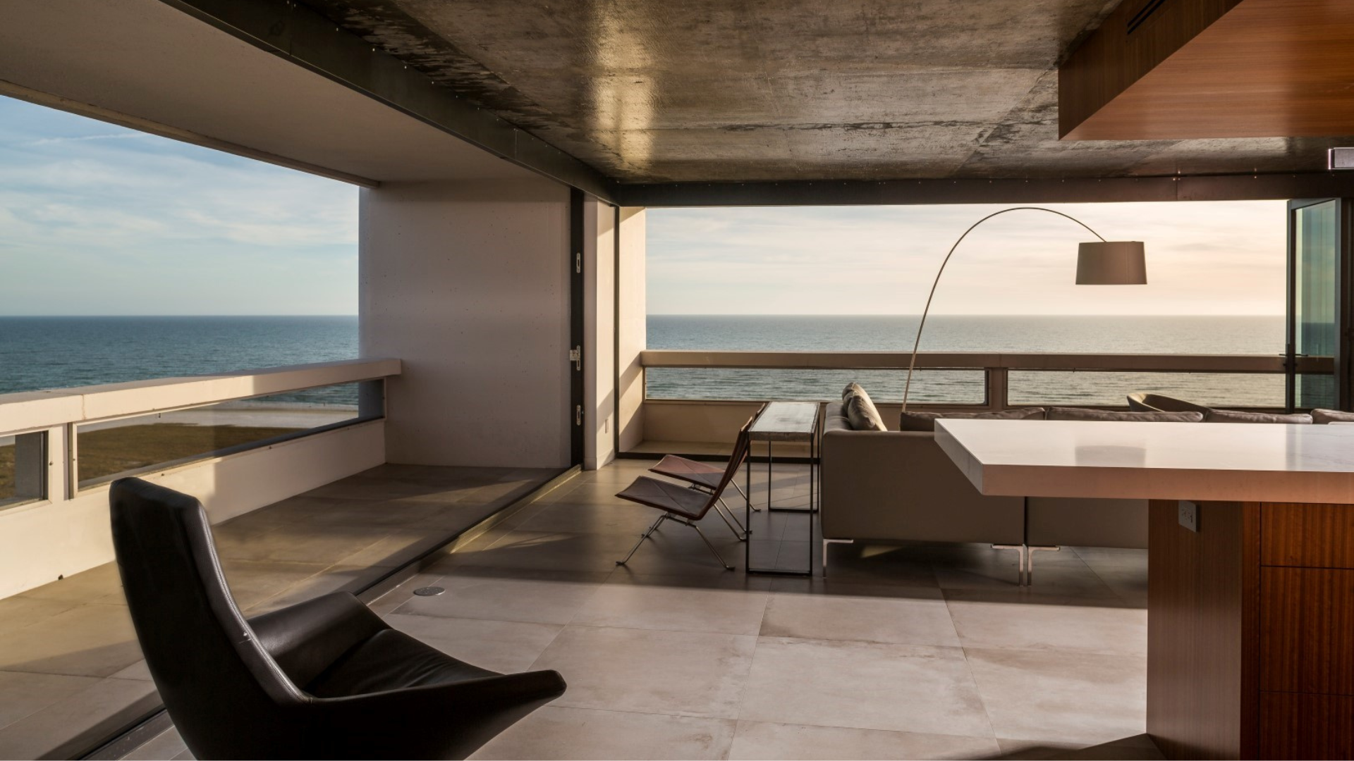 Modern living room with large windows and balcony overlooking the ocean. Minimalist furniture includes a sofa, desk, lamp, and chairs. Warm natural light fills the space.