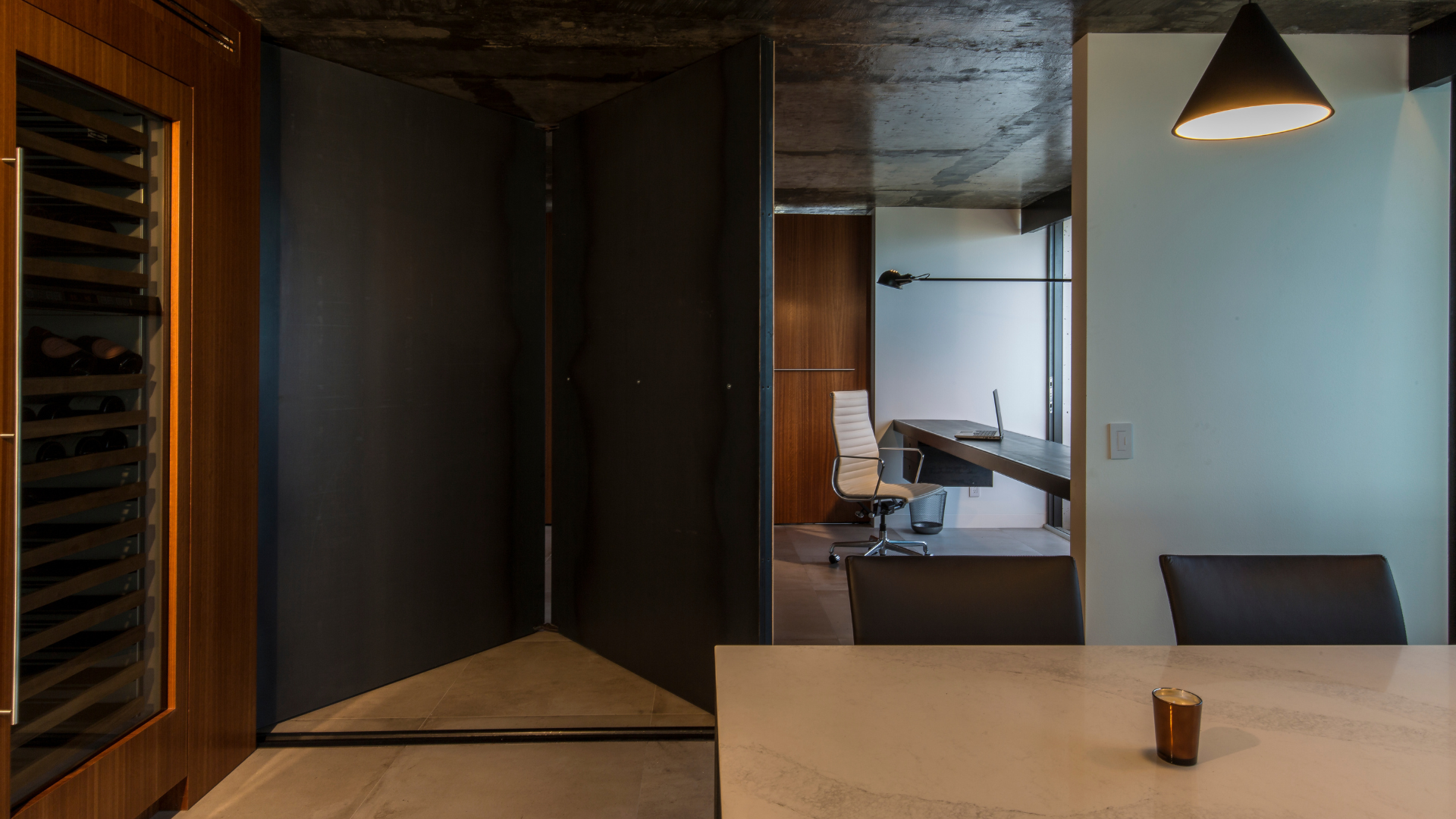 A modern office space with a white chair at a desk, seen through a large black folding partition. A marble table with a small cup, a black chair, and a hanging lamp are in the foreground.