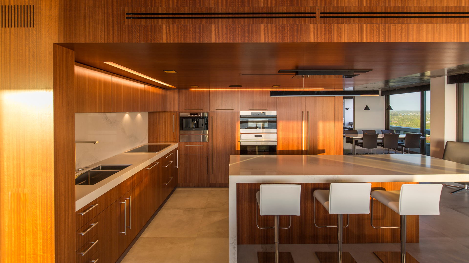 Modern kitchen with wood paneling, built-in appliances, white countertops, and three white bar stools at an island. Sunlight shines through large windows, revealing a dining area in the background.