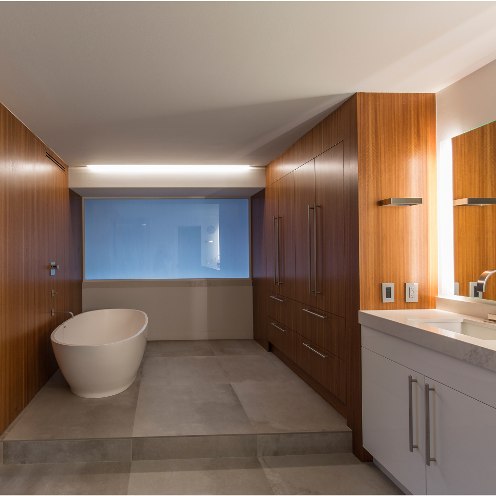 Modern bathroom with a freestanding white bathtub, large wood-paneled cabinets, a wide mirror, and a white countertop sink, illuminated by sleek overhead lighting.