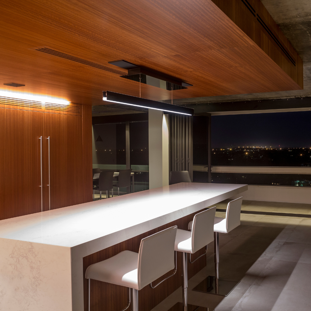 Modern kitchen with a long white island, three white chairs, wood paneling, minimalist lighting, and large windows showing a night cityscape in the background.