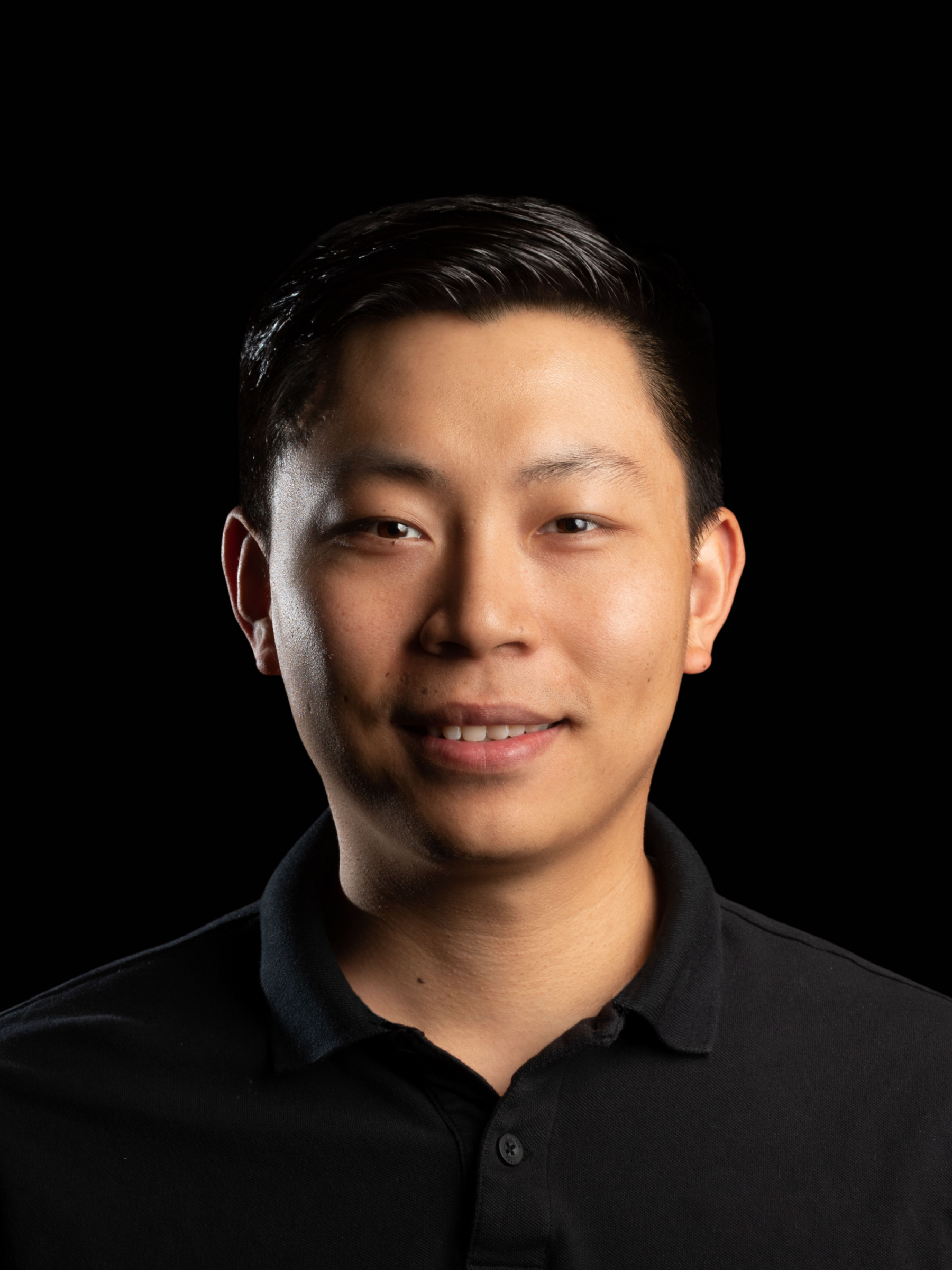 A young man with short black hair, wearing a black polo shirt, smiles softly against a solid black background.