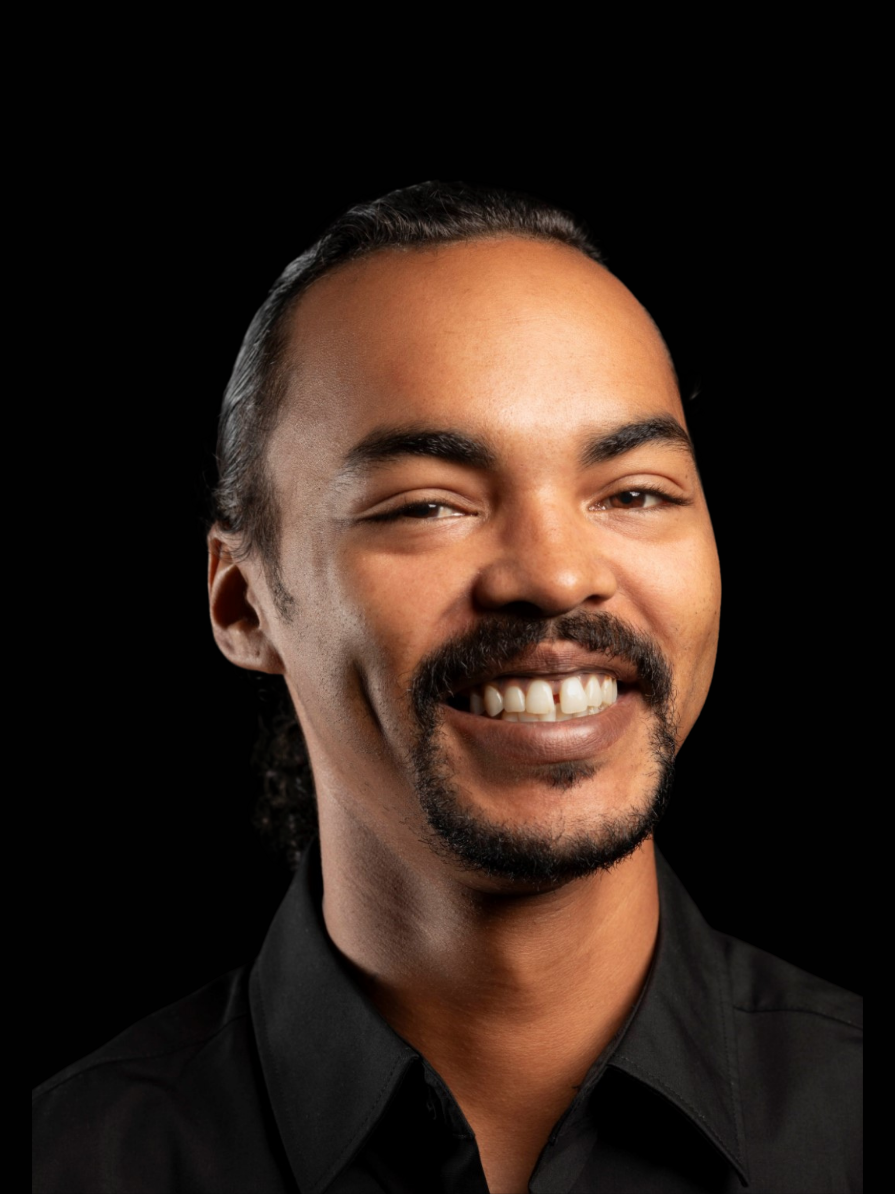 A smiling man with a mustache and goatee, wearing a black collared shirt, is shown against a solid black background.