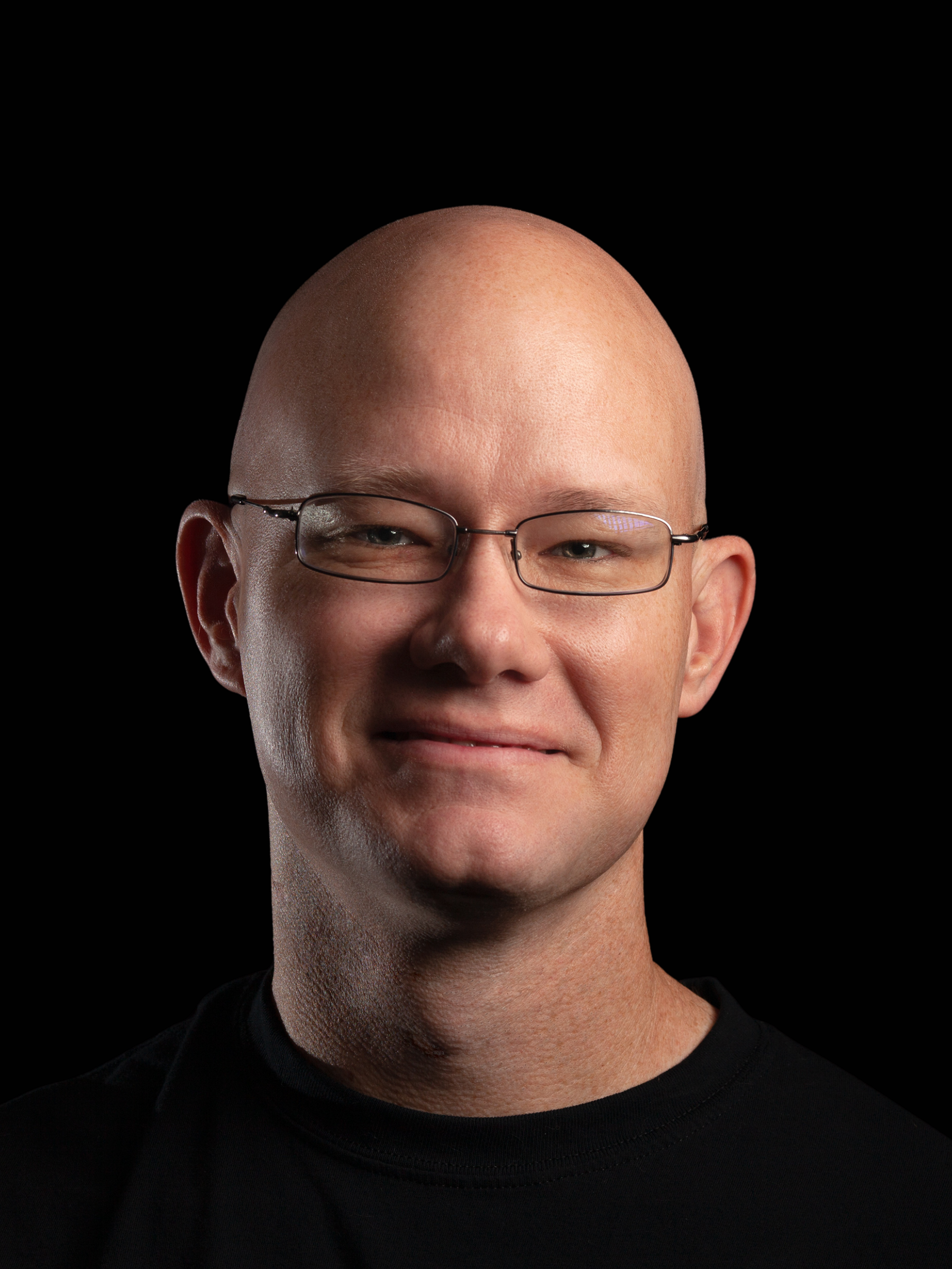 A bald person with glasses and a slight smile, wearing a black shirt, is shown against a plain black background.