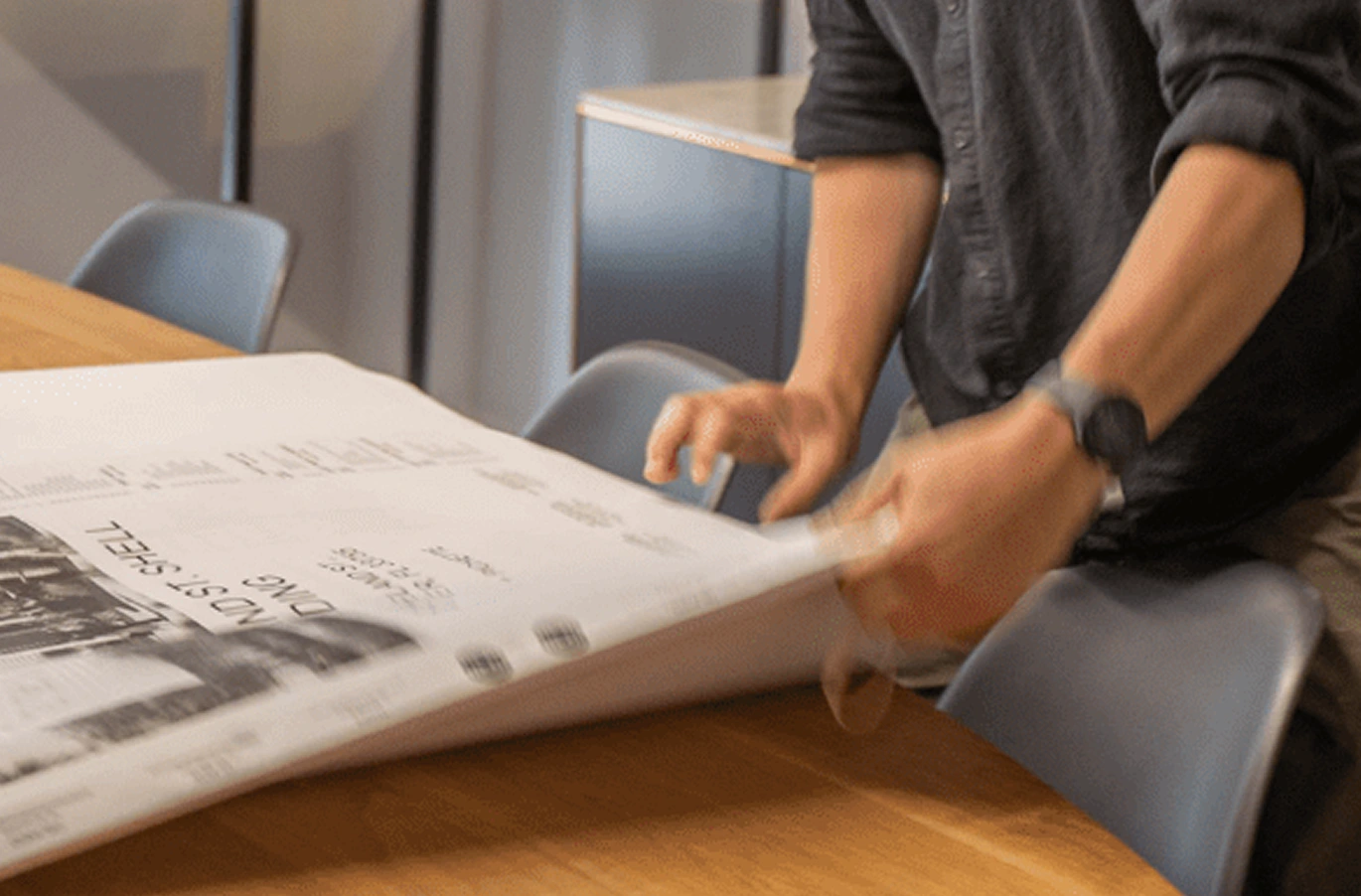 A person in a dark shirt is spreading a large Halflants + Pichette sheet with text and images across a wooden table, surrounded by several chairs.