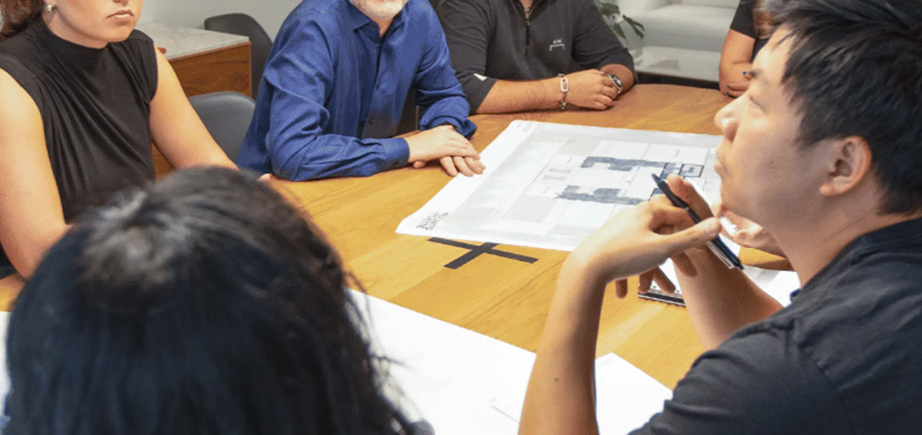 Four people sit around a wooden table with architectural blueprints, engaged in an Architect-Led Design-Build discussion. One holds a pen while another in a blue shirt collaborates in this dynamic work session.