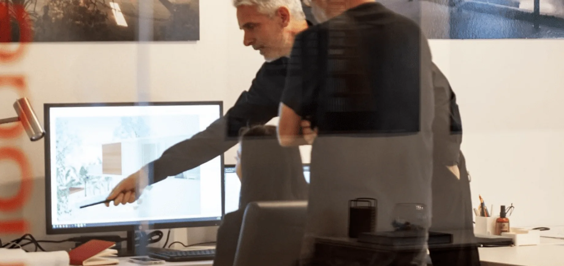 Two people in an office, one pointing at a computer screen while the other stands nearby—an Architect-Led Design-Build meeting viewed through glass, with reflections and a slightly blurred effect.