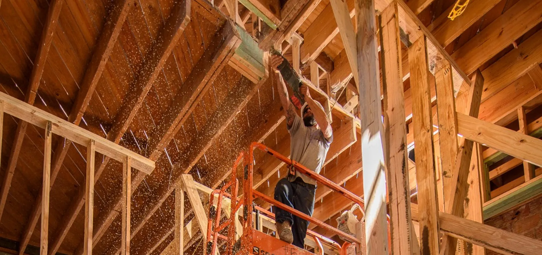 A construction worker on a lift uses a power saw to cut a wooden beam in an Architect-Led Design-Build project, with sawdust flying as sunlight filters through the unfinished structure.