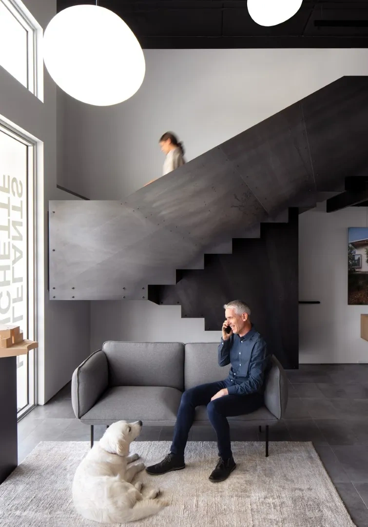 A man sits on a gray sofa talking on the phone, with a white dog on the rug nearby. A person walks upstairs in a modern Halflants + Pichette room with large round lights.