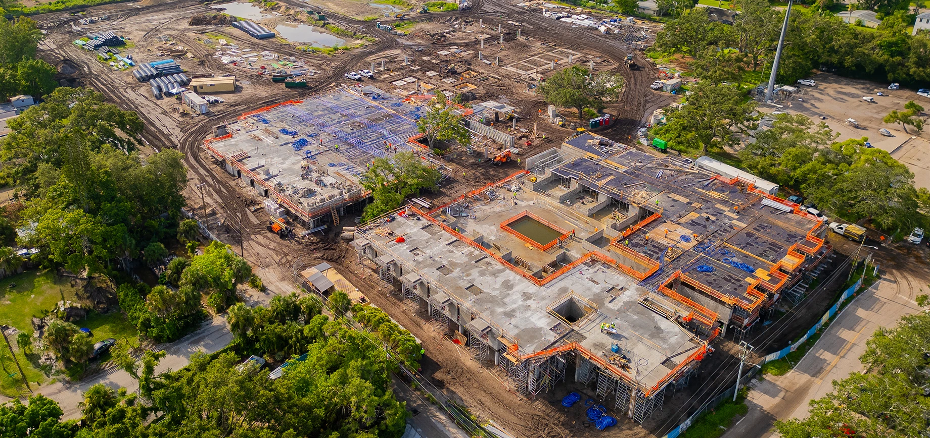 Aerial view of a large commercial architecture construction site with multiple concrete buildings in progress, surrounded by dirt, trees, and construction materials.