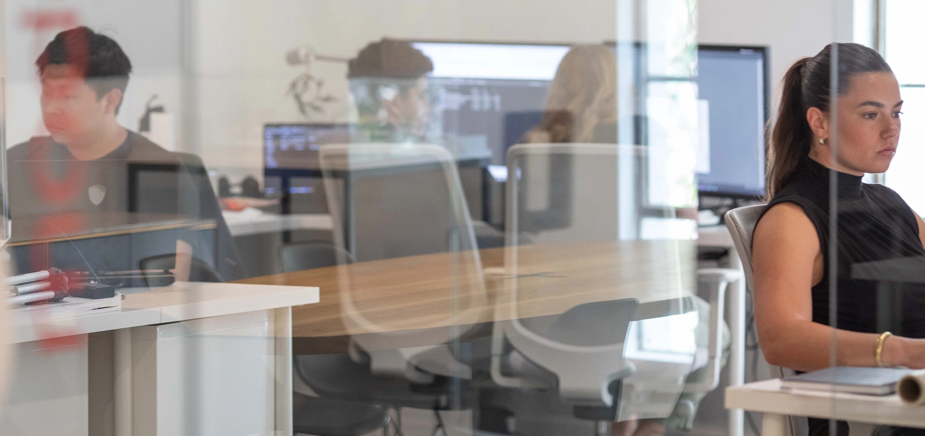Four people work at desks with computers in a modern office space defined by sleek commercial architecture. Glass walls and reflections create a layered look, while one woman in the foreground faces right as she works.