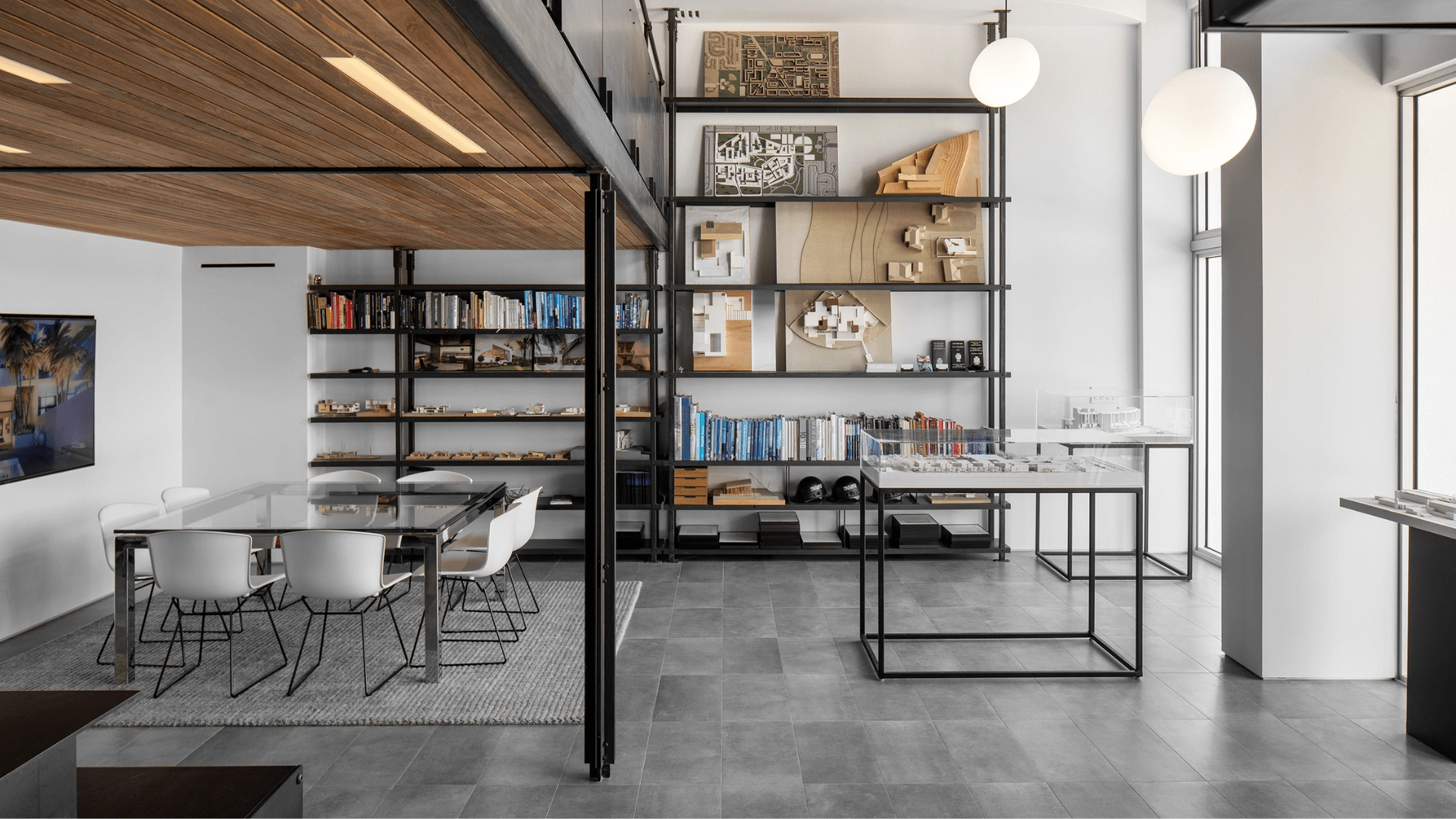 Modern office space by Halflants + Pichette with a glass table and white chairs, industrial shelves holding books and architectural models, large windows, gray tiled flooring, and hanging globe lights.