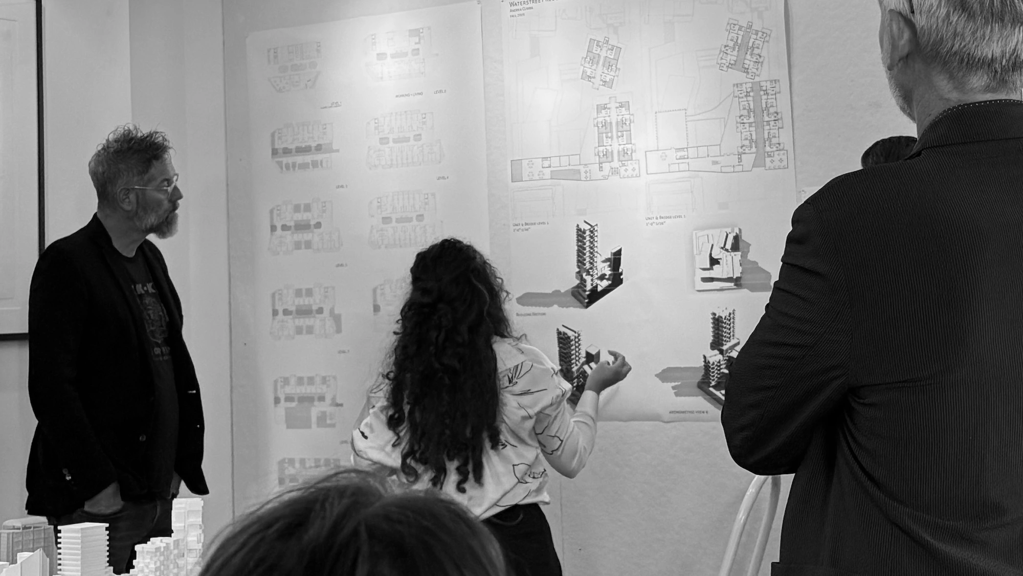 A woman presents architectural plans and models to a small group at USF. Three-dimensional building models and technical drawings are displayed on the wall.