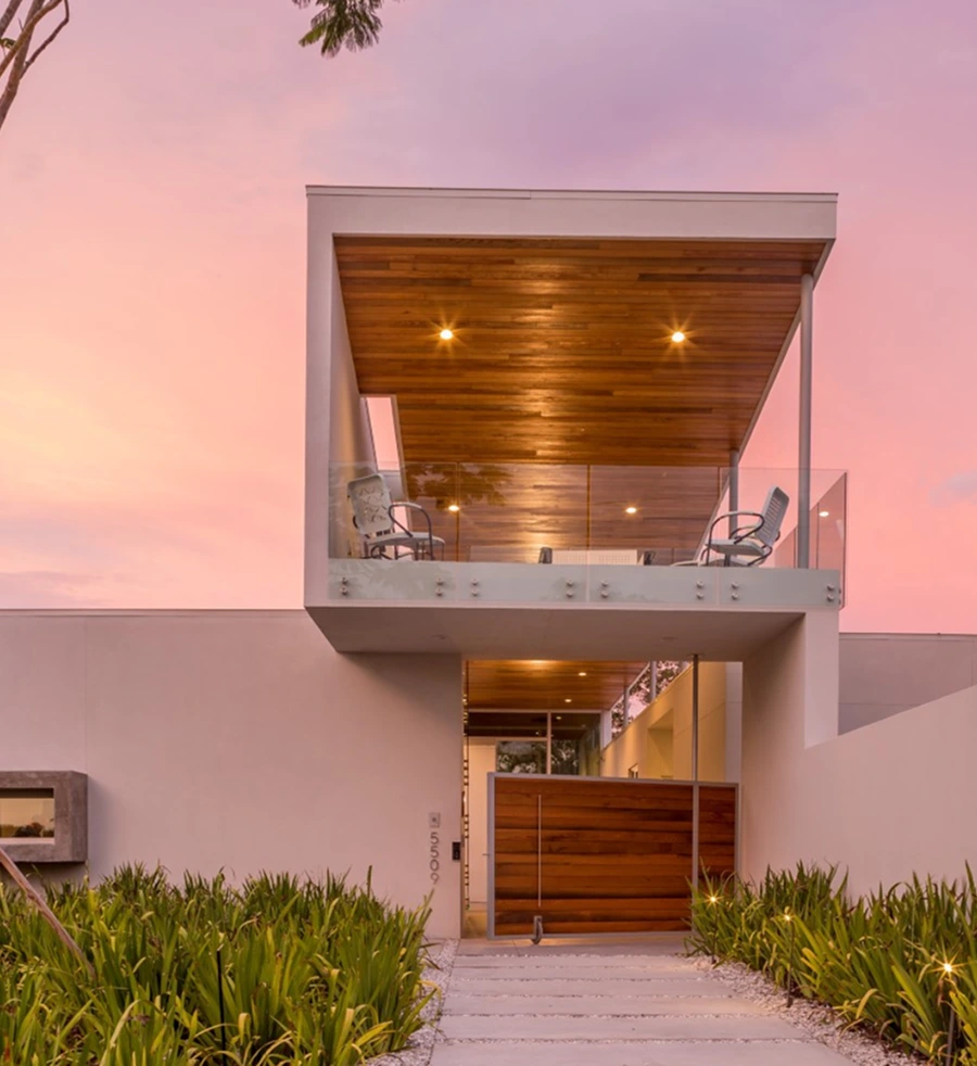 Modern Halflants + Pichette house with a white facade, wooden accents, balcony seating, glass railings, and lush green plants at the entrance, set against a pink sunset sky.
