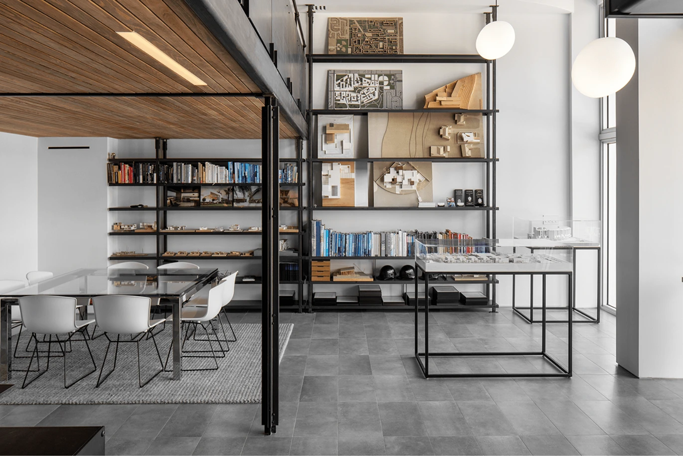Modern office with glass table, white chairs, large shelving holding books and architectural models, and display tables with scale models, all in a bright, minimalist space with grey floors and large windows.