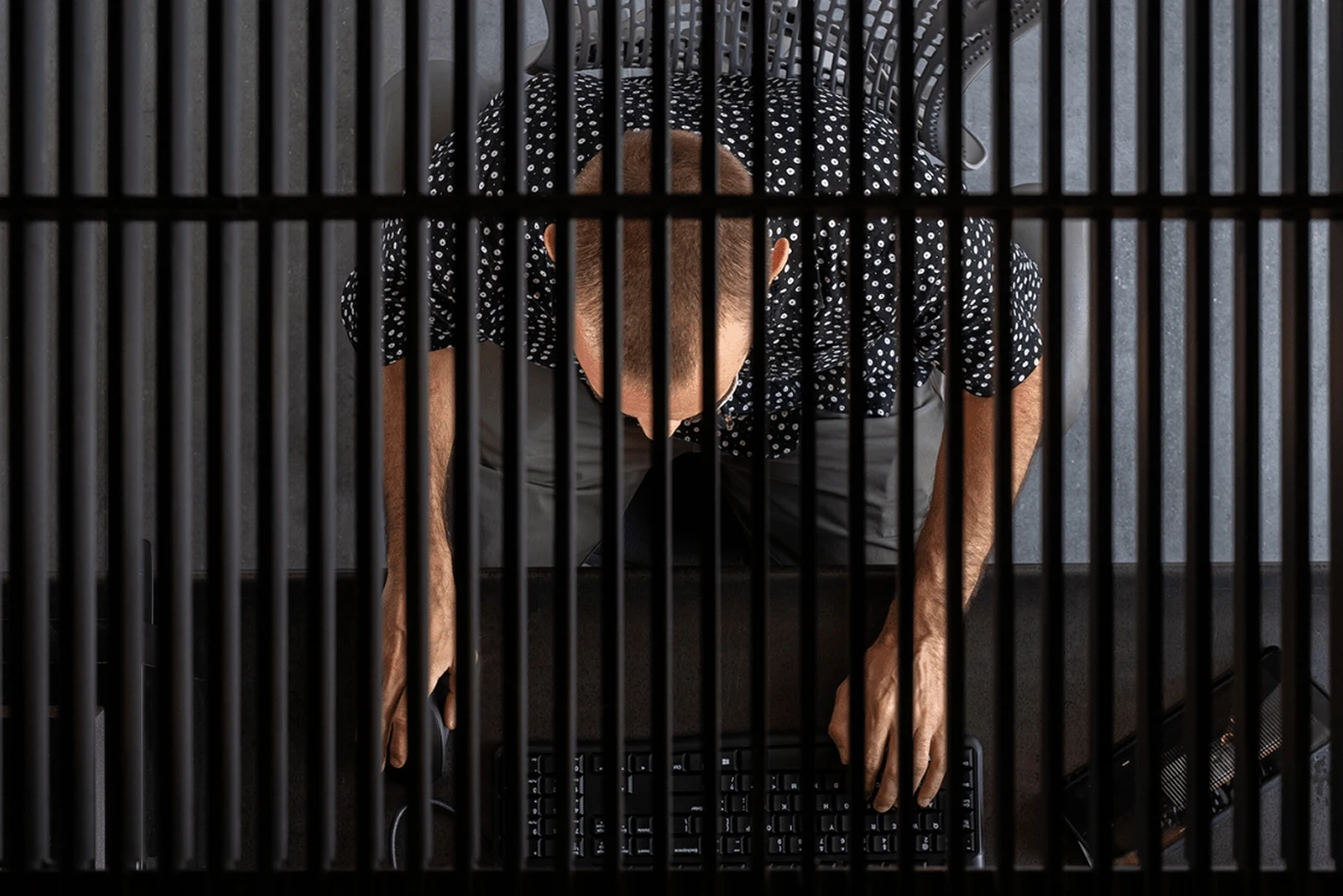 Top view of a person sitting at a desk, typing on a keyboard, seen through vertical black bars—a striking visual echoing themes of confinement in modern commercial architecture.