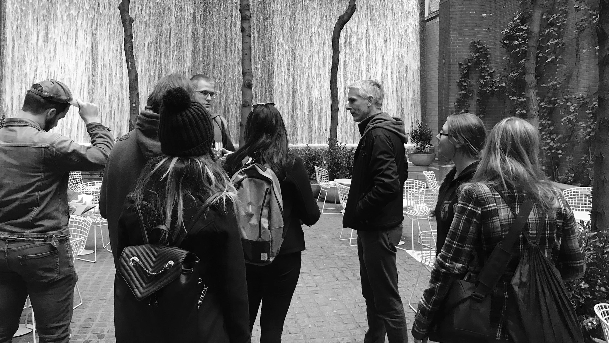 A group of people stands in an outdoor courtyard at USF, facing a tall wall with cascading water. Some wear backpacks and hats. Empty wire chairs and tables are scattered around. The scene is in black and white.